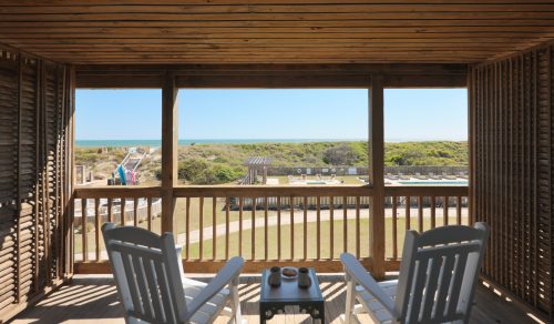 Two white rocking chairs on a wooden balcony overlook grassy dunes, pool, and distant blue ocean