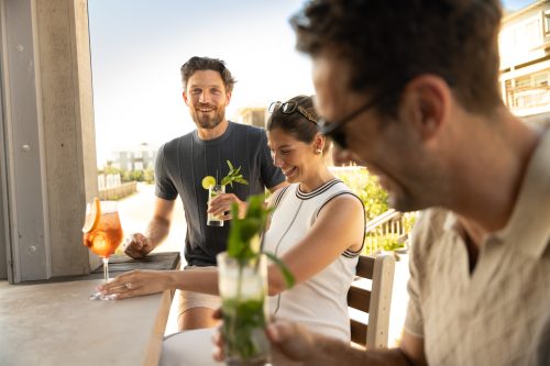 Three friends relax outdoors, drinking cocktails at a sunlit bar or patio with casual summer atmosphere.