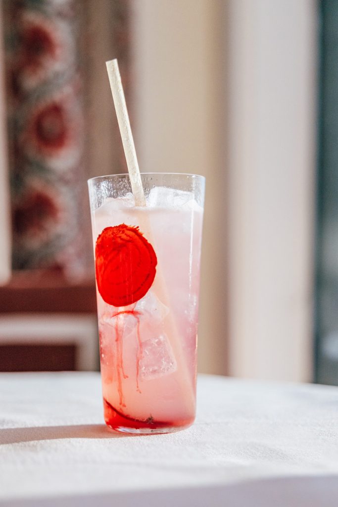 Tall glass filled with pink iced drink, large red garnish, and straw on a white tablecloth.