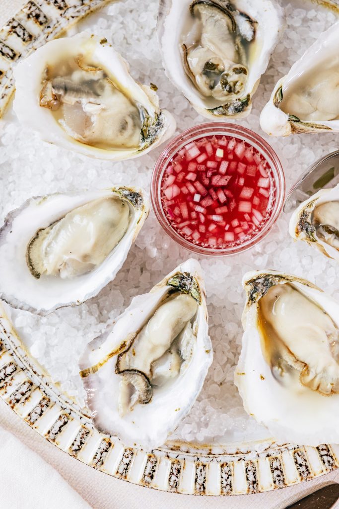 Plate of fresh oysters on ice served with a small dish of red mignonette sauce.