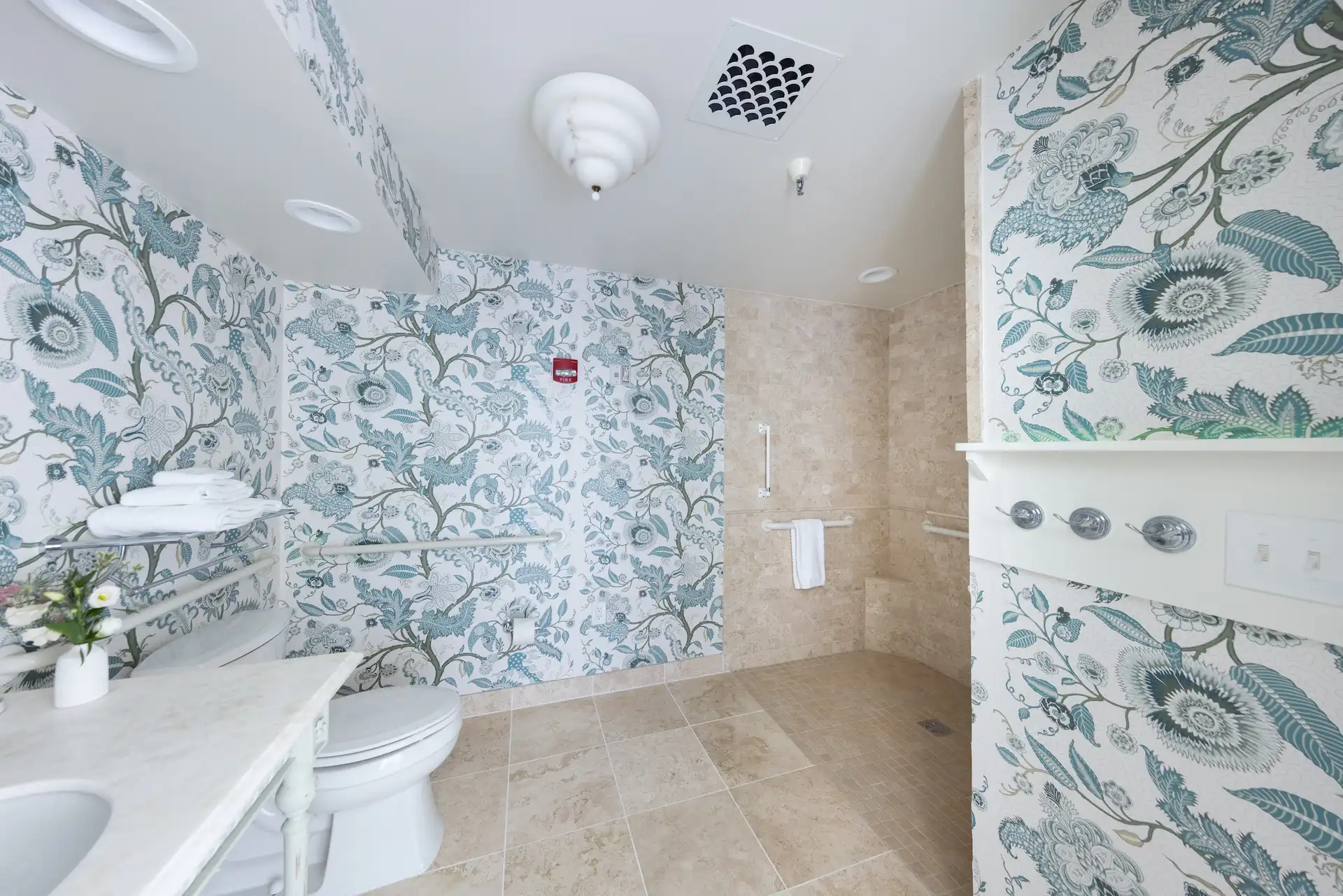 Bathroom with bold floral wallpaper, marble sink, and beige tiled shower blending coastal charm with eclectic elegance.