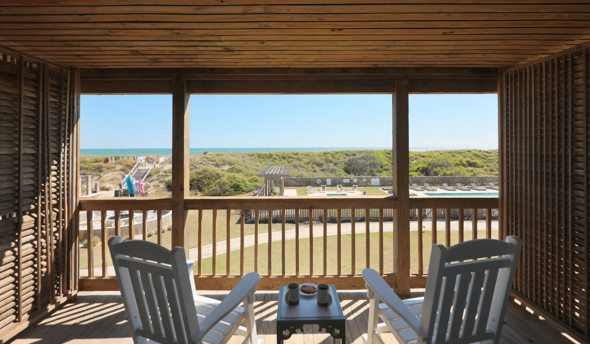 White rocking chairs on a wooden balcony facing the ocean, with a small table and coastal path framed by lush greenery and blue sky.