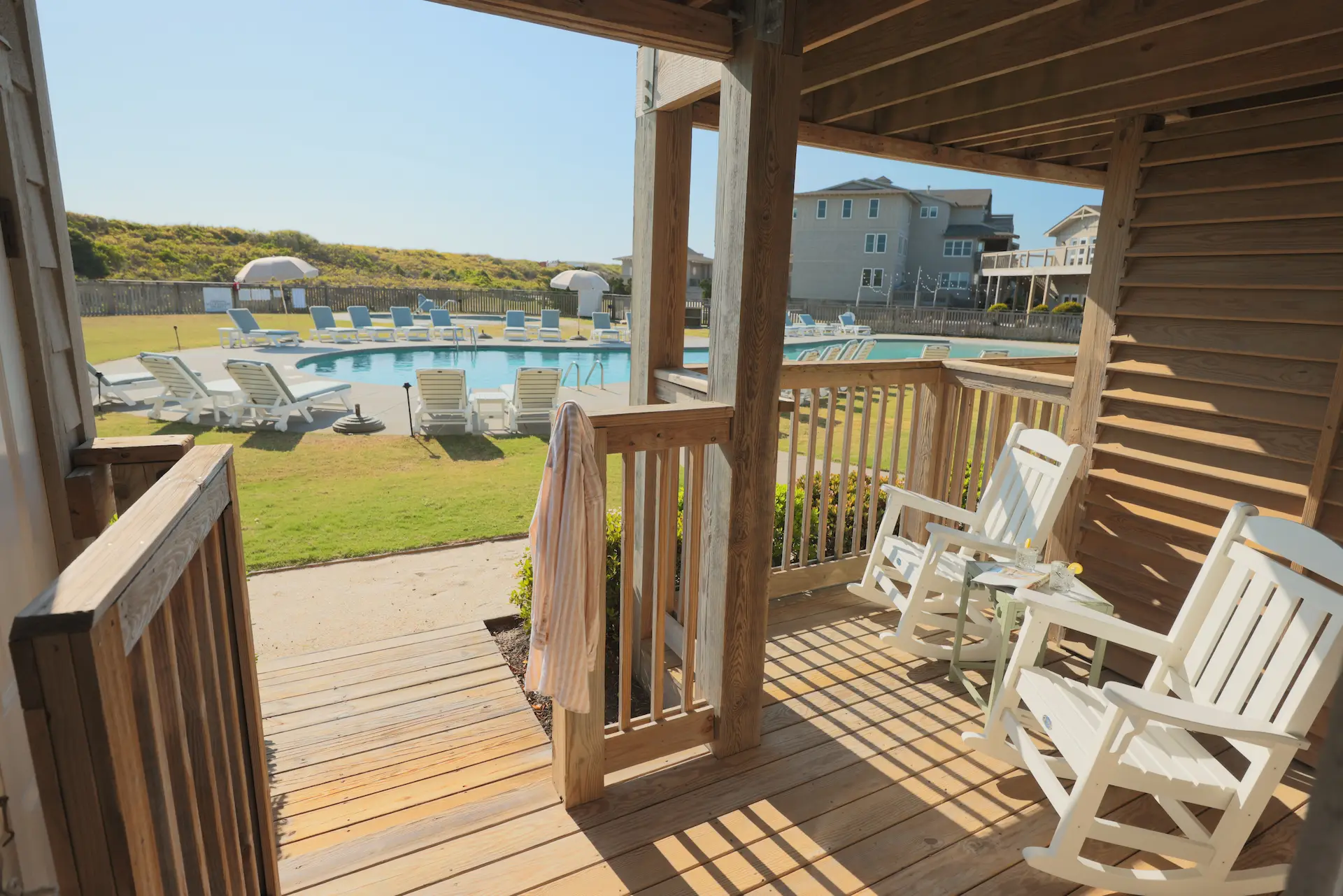 White rocking chairs on a porch overlooking a sunny pool area with lounge chairs, umbrellas, and a relaxed resort vibe.