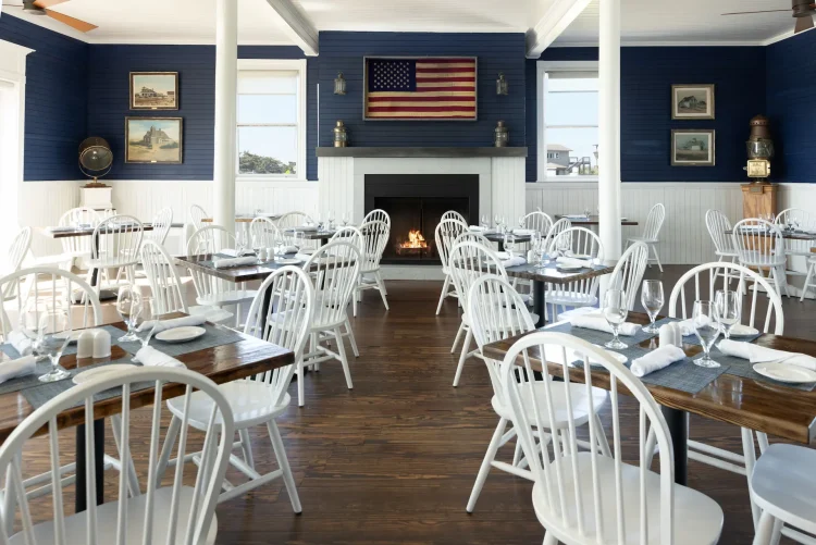 Coastal-style dining room with white chairs, dark wood tables, fireplace, and American flag decor.
