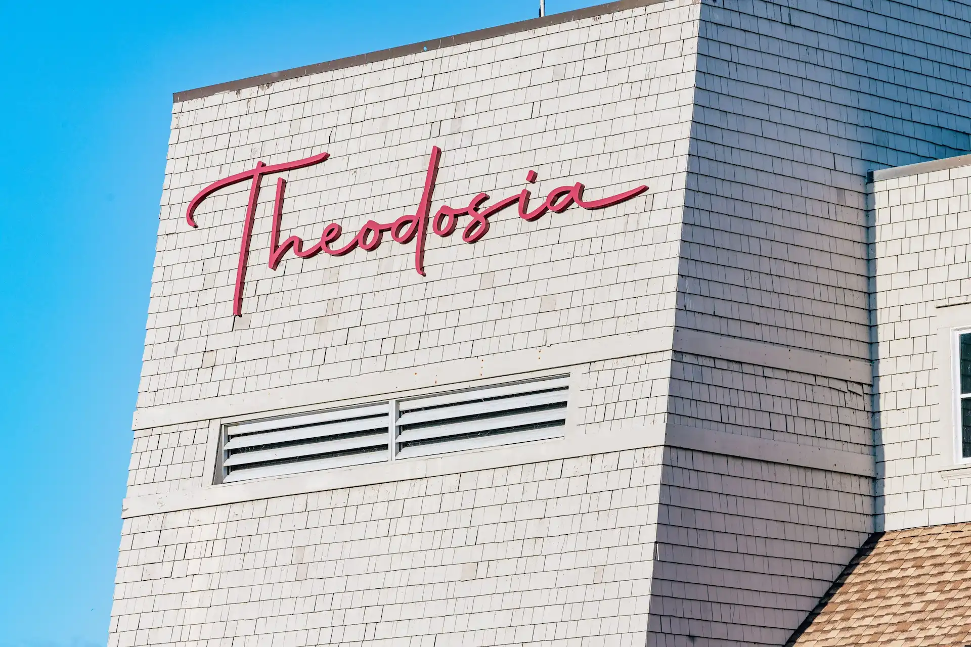Exterior of white shingle-style building with red cursive “Theodosia” lettering, set against a clear blue coastal sky.