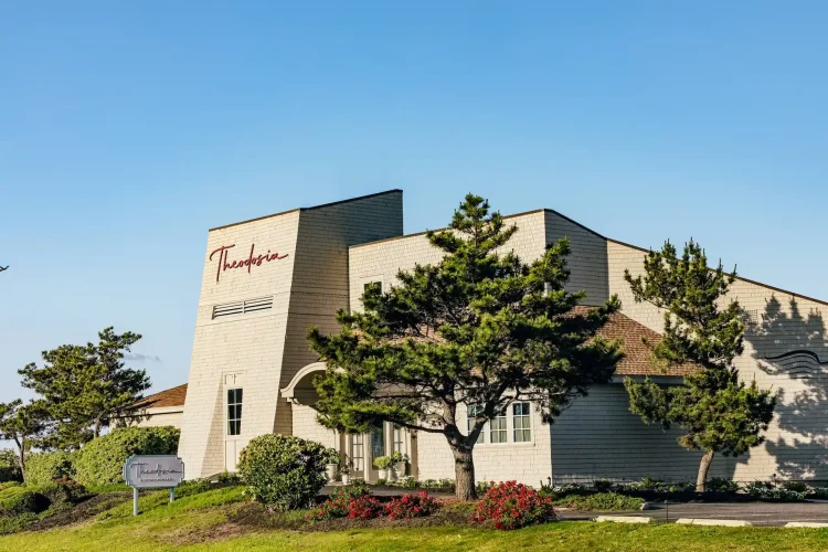 Exterior of white shingle-style building with red cursive Theodosia lettering, set against a clear blue coastal sky.