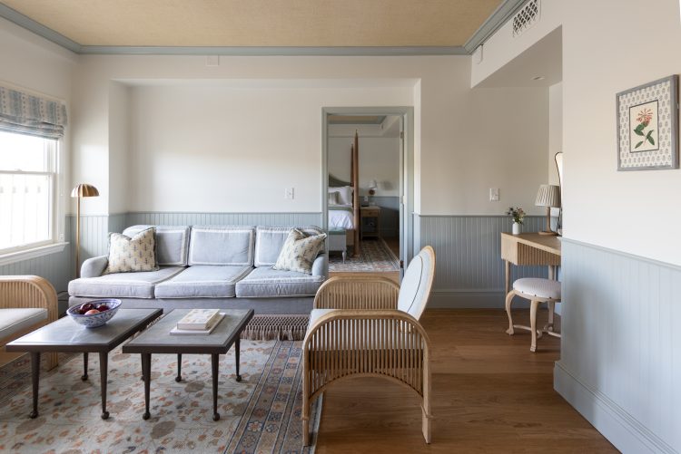 Bright living room with gray couch, wicker chairs, nested tables, rug, bedroom visible through doorway.