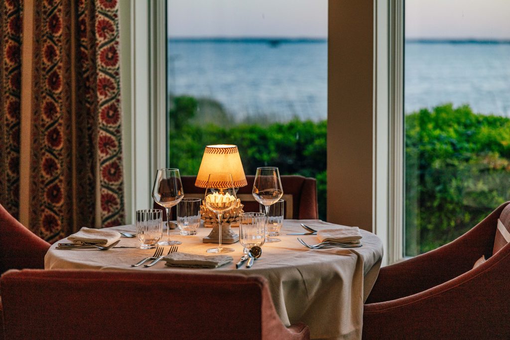 Elegant seaside dining table with soft lamp lighting, wine glasses, and ocean view at dusk.