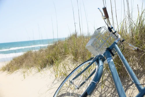 Blue bicycle with basket resting on sandy beach near ocean waves and tall dune grass.
