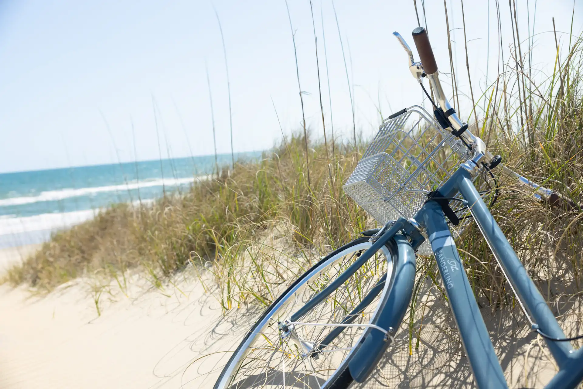 Blue bicycle with basket resting on sandy beach near ocean waves and tall dune grass.