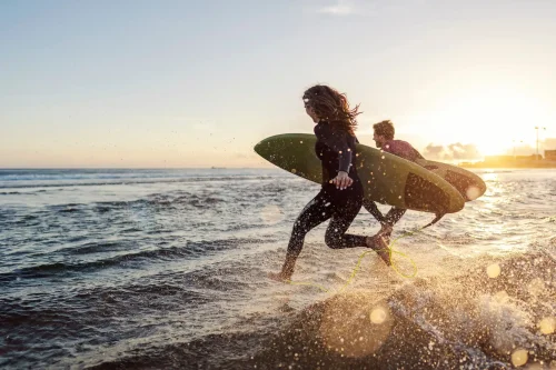 Two surfers in wetsuits run into the ocean at sunrise, boards in hand, golden light casting energy across the calm waves.