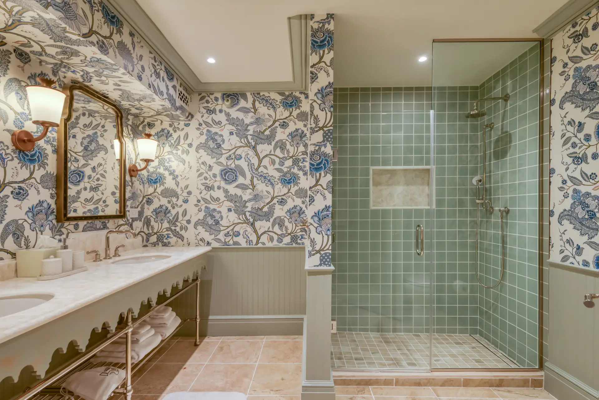 Elegant bathroom with floral wallpaper, double sink vanity, gold mirrors, and glass-enclosed tiled shower.