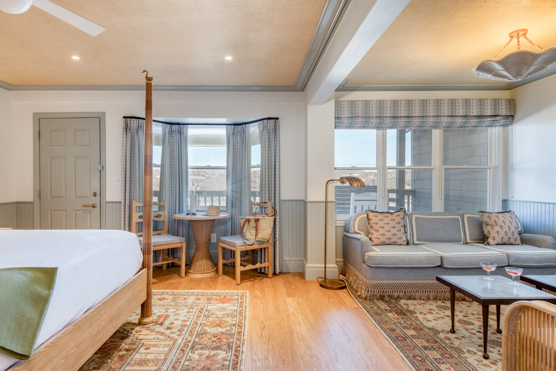 Bright coastal suite with four-poster bed, cozy sitting area, bay window, and patterned rugs.