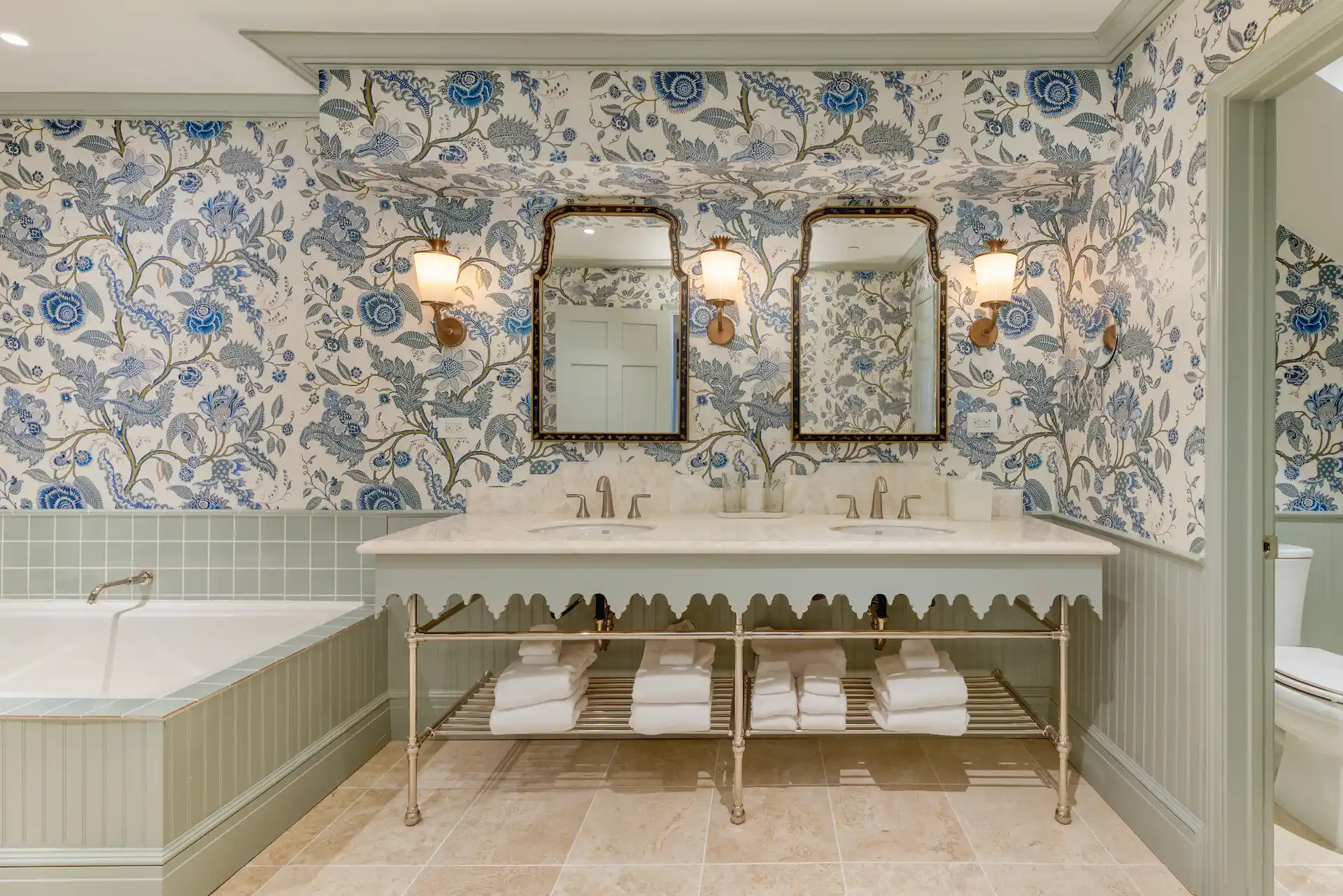 Elegant bathroom with double sinks, mirrors, blue floral wallpaper, soft lights, and neatly stacked towels.
