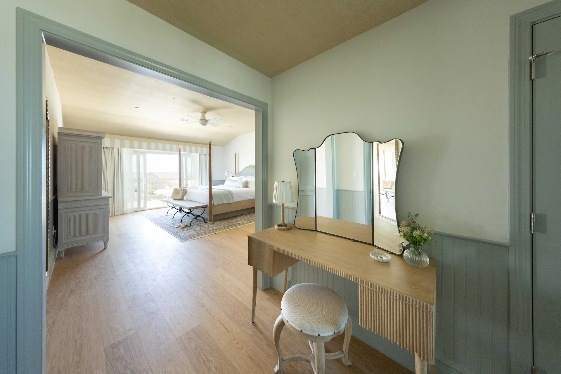 Bright coastal bedroom with vanity area, four-poster bed, wooden floors, and sunlight streaming through windows.