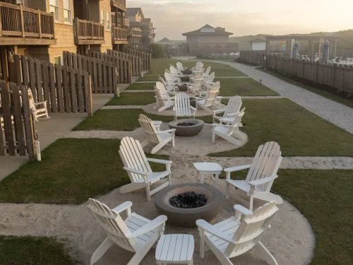 Outdoor seating area with white Adirondack chairs arranged around fire pits between beachfront cottages.