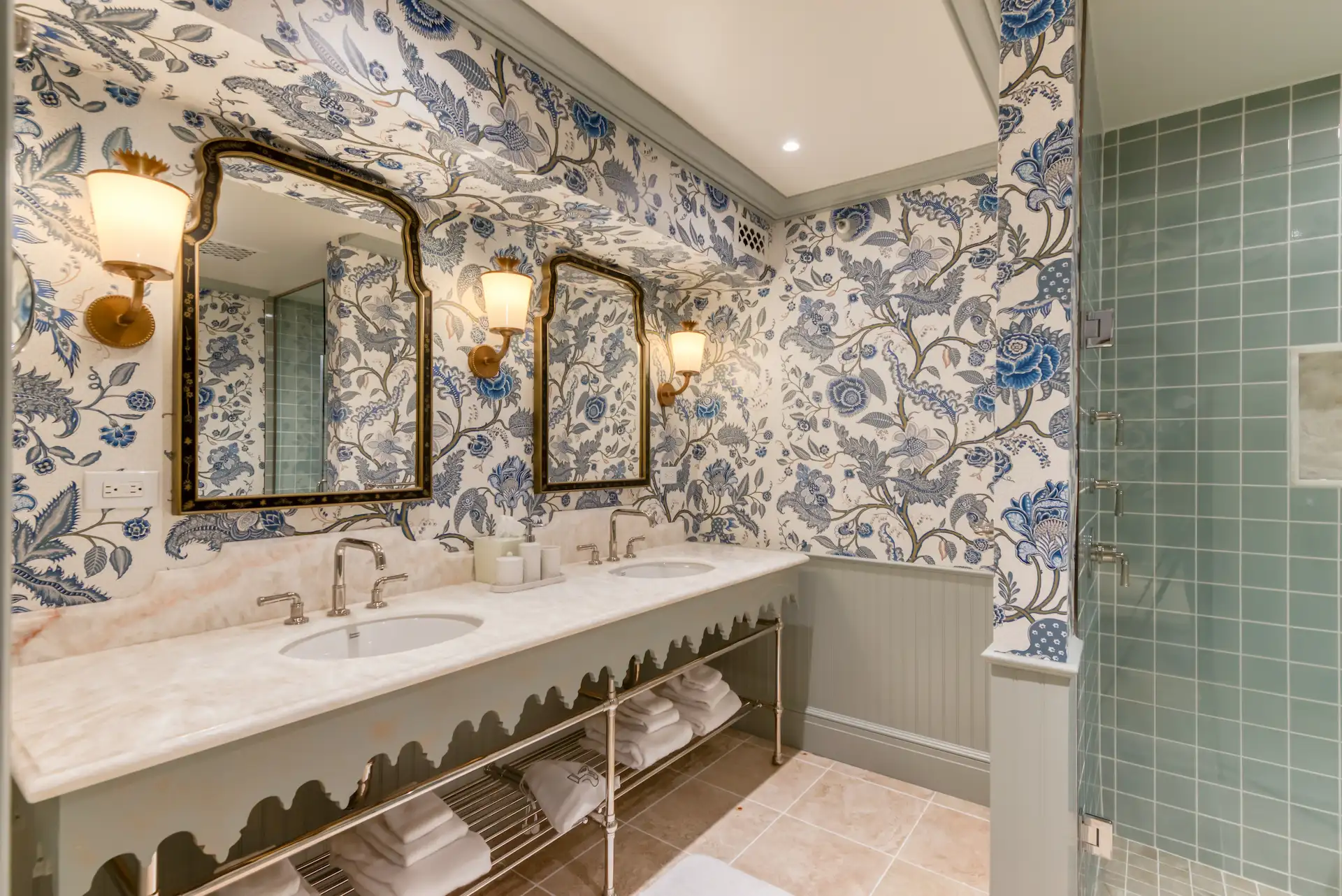 Elegant bathroom with floral blue wallpaper, double vanity, brass mirrors, and glass-tiled shower area.
