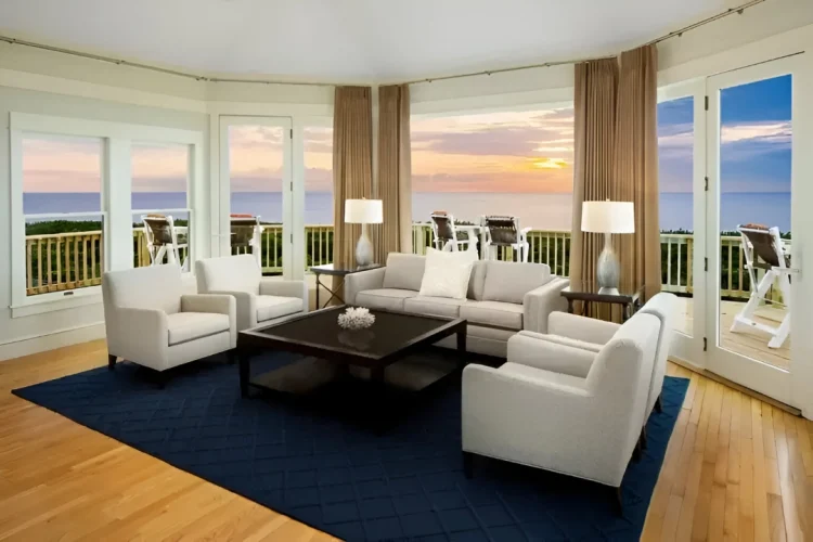 A serene living room with large windows overlooking the sea, bathed in the golden-orange glow of a breathtaking sunset—where time seems to slow down.