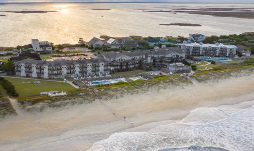 Aerial view of beachfront resort with pools, buildings, and ocean at sunset.