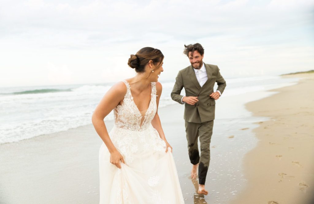 Bride and groom joyfully run barefoot along the beach under a bright blue sky.
