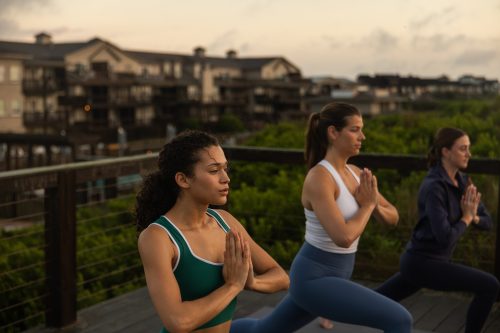 Three people practicing yoga outdoors at sunrise, hands in prayer pose, with wooden buildings behind.