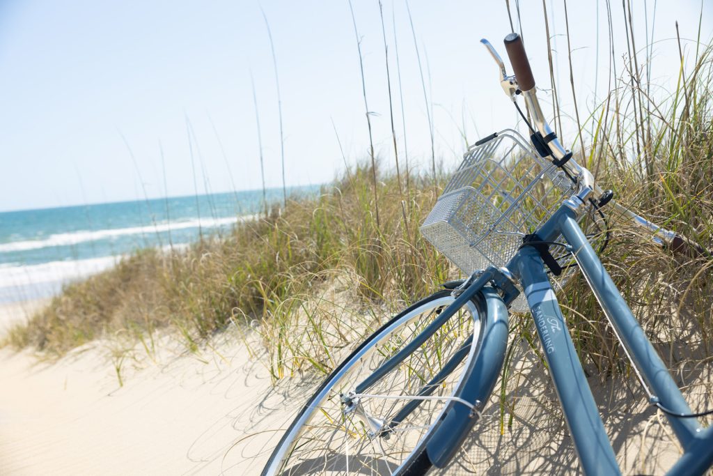Blue bicycle with basket resting on sandy beach near ocean waves and tall dune grass.