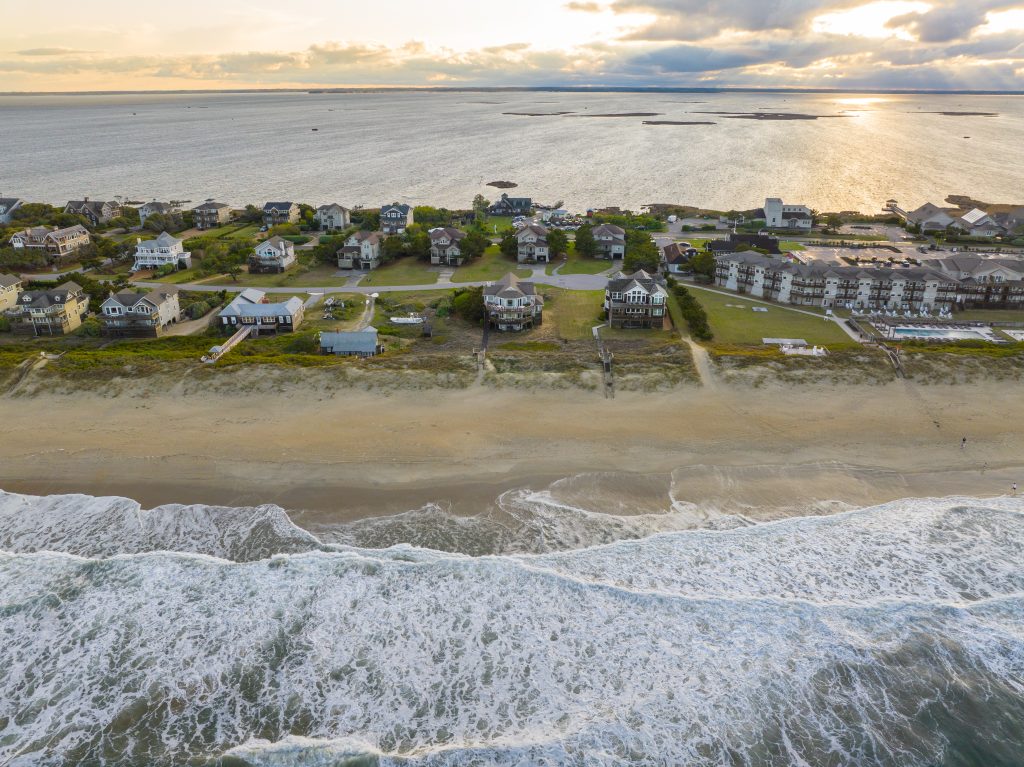 Aerial view of beachfront homes along the coastline with waves crashing under a cloudy sunset.