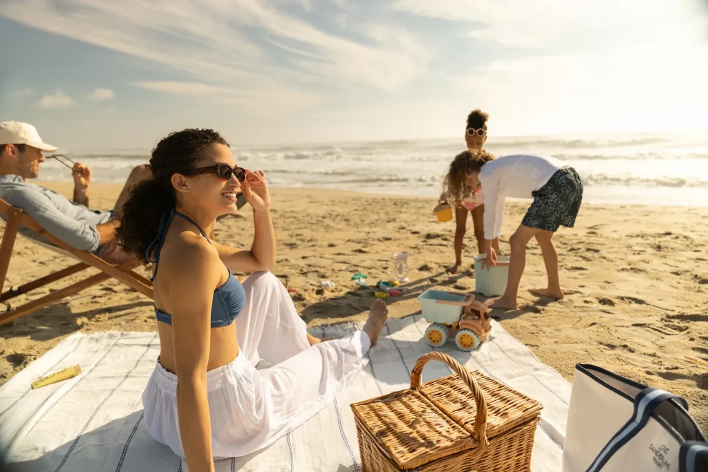Smiling family enjoying sunny beach picnic while children play in sand near ocean waves.