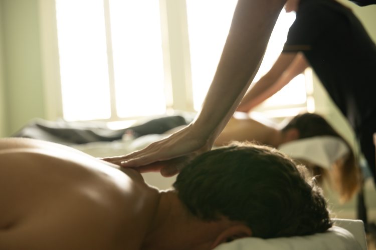 Two people receiving relaxing back massages in a sunlit spa room, therapists working by window light.