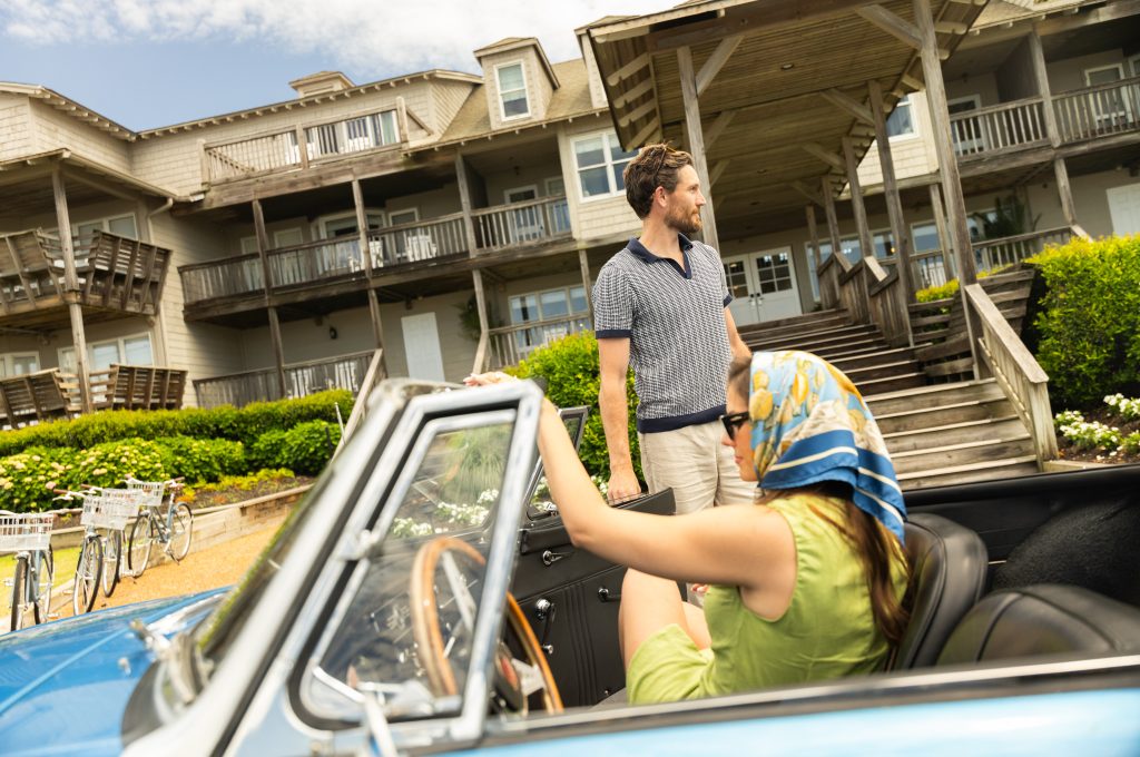 Couple with vintage convertible parked outside seaside inn, man standing as woman sits adjusting scarf.