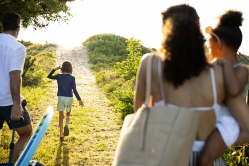 Group walking along sandy path toward the beach, with surfboard, tote bag, and children enjoying the sunny coastal greenery.