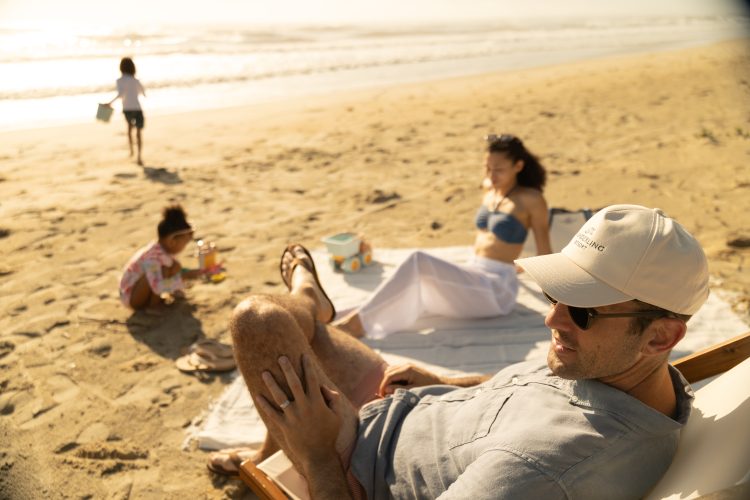 Family relaxing on sandy beach, man reclining in chair while children play near shoreline.