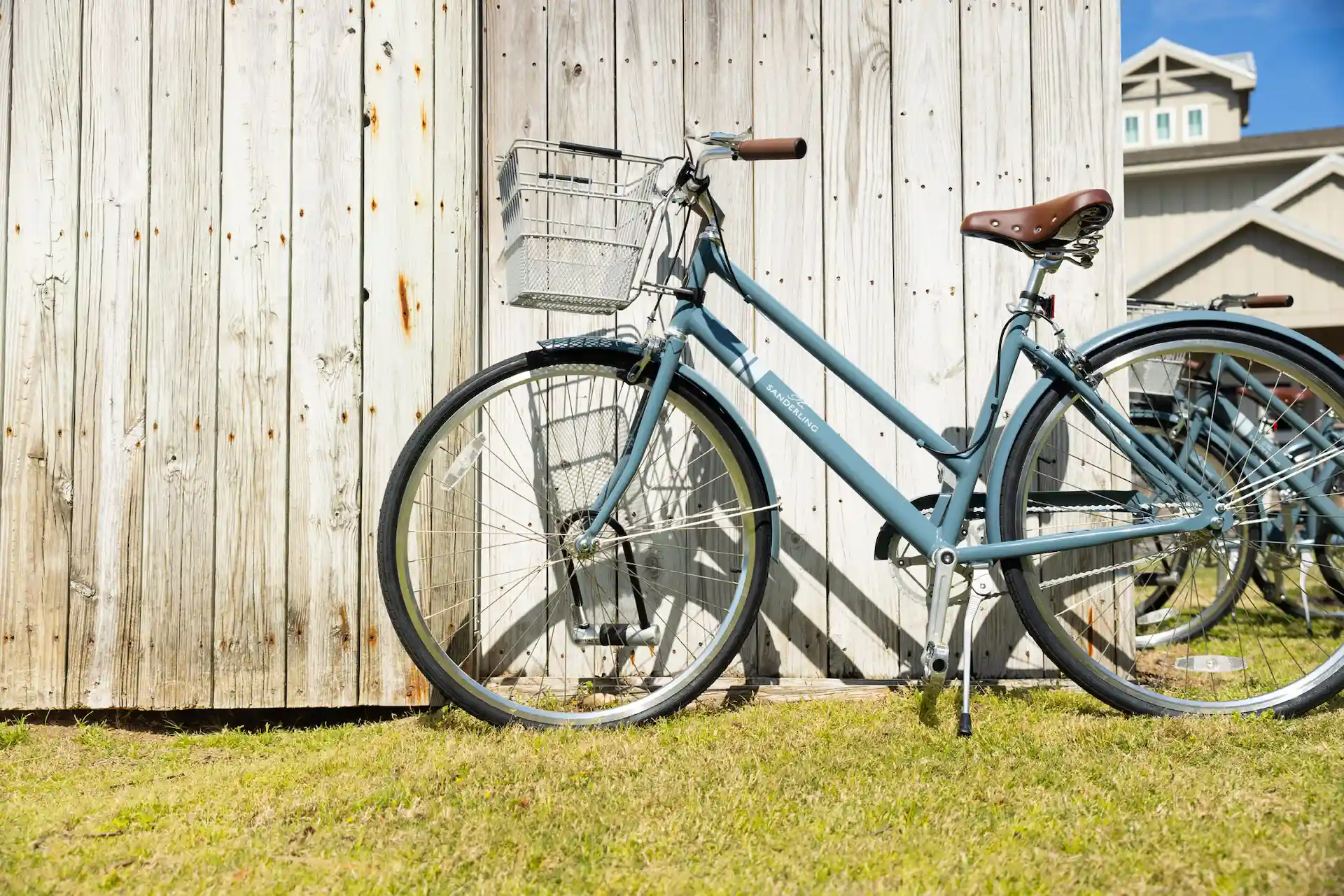 lowRes_tSr_Bikes_IMG_1745 Blue bicycle with basket parked on grass beside rustic wooden shed in sunny coastal setting.