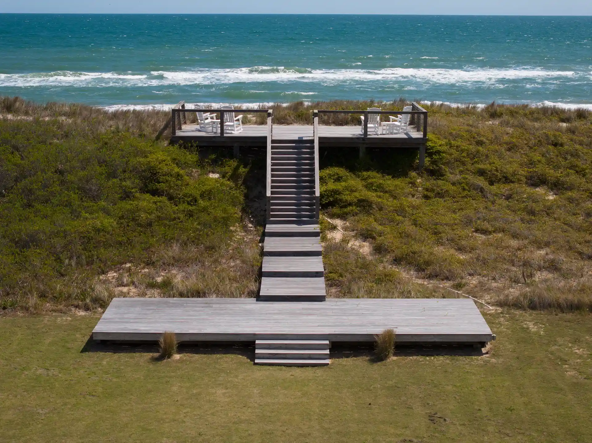 Wooden boardwalk and stairs lead to ocean-view deck with white chairs among coastal dunes.