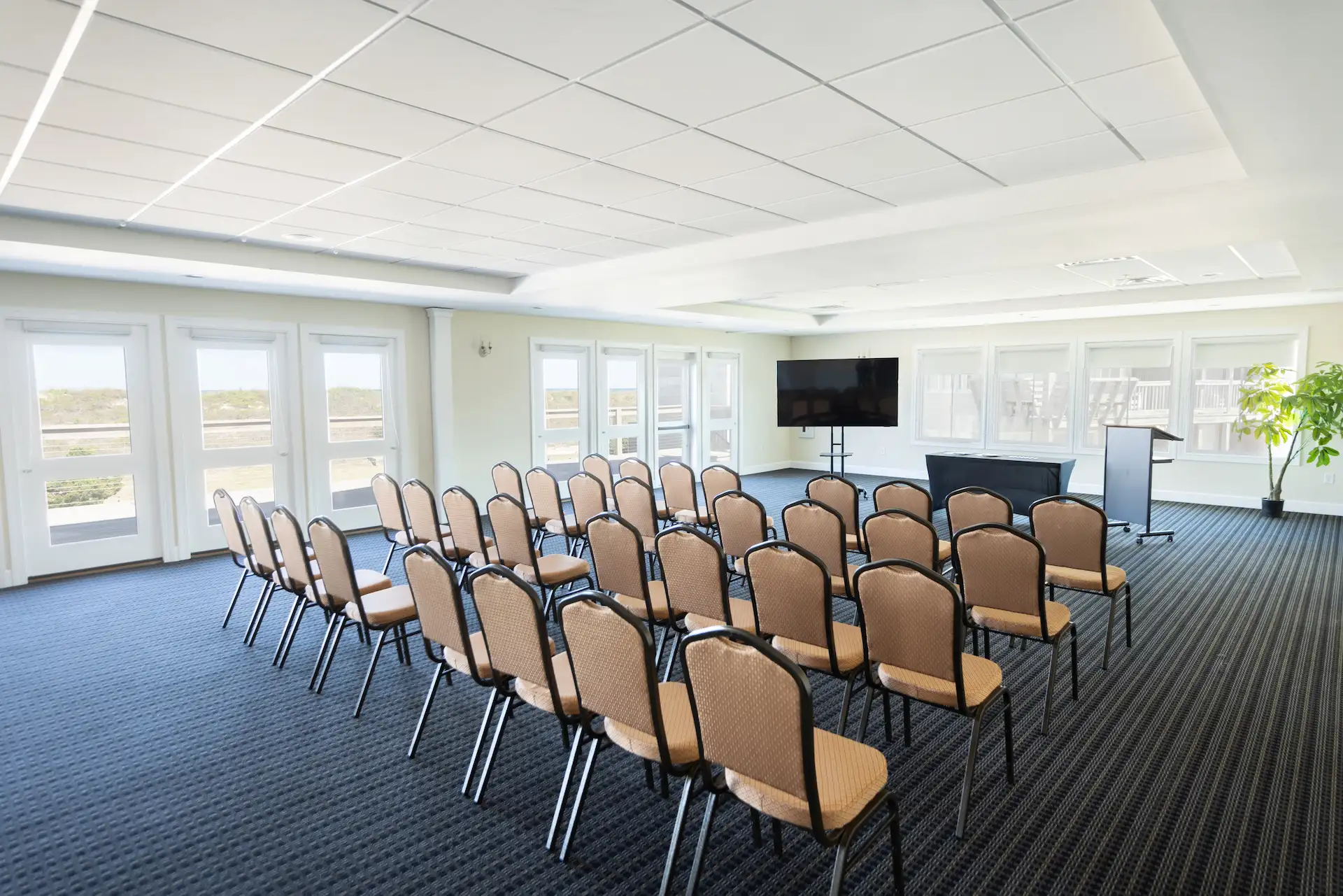 Modern meeting room with rows of chairs facing podium and screen, lit by natural light from large windows.