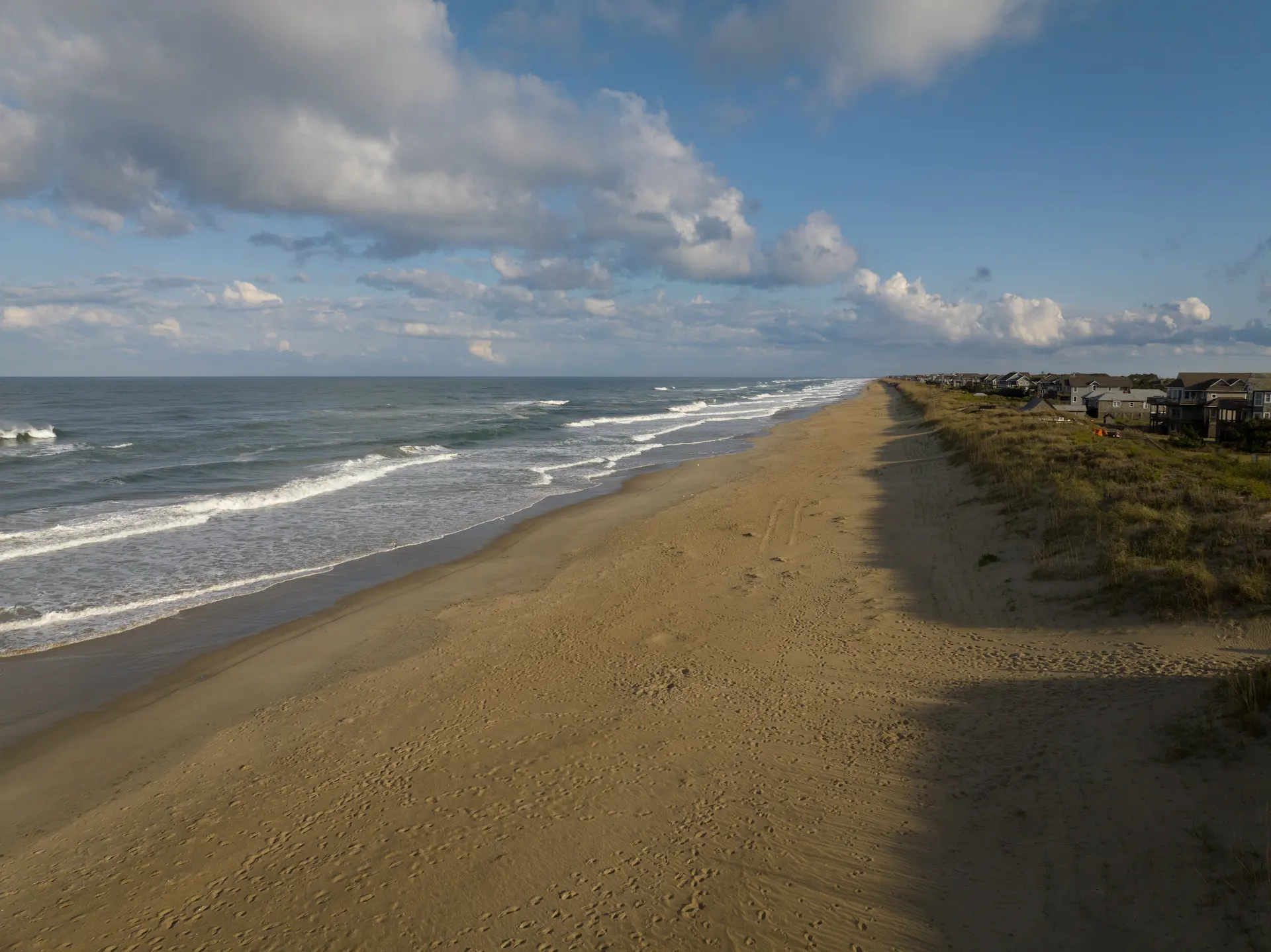 Wide sandy beach near The Sanderling hotel, with gentle waves and coastal homes under a partly cloudy sky.