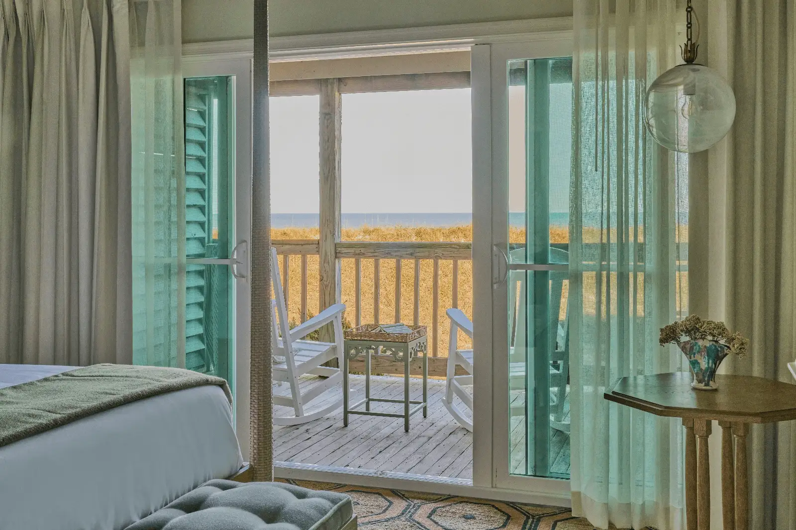 Coastal bedroom with mint curtains and ocean-view balcony, featuring white linens, rocking chairs, and a peaceful vibe.