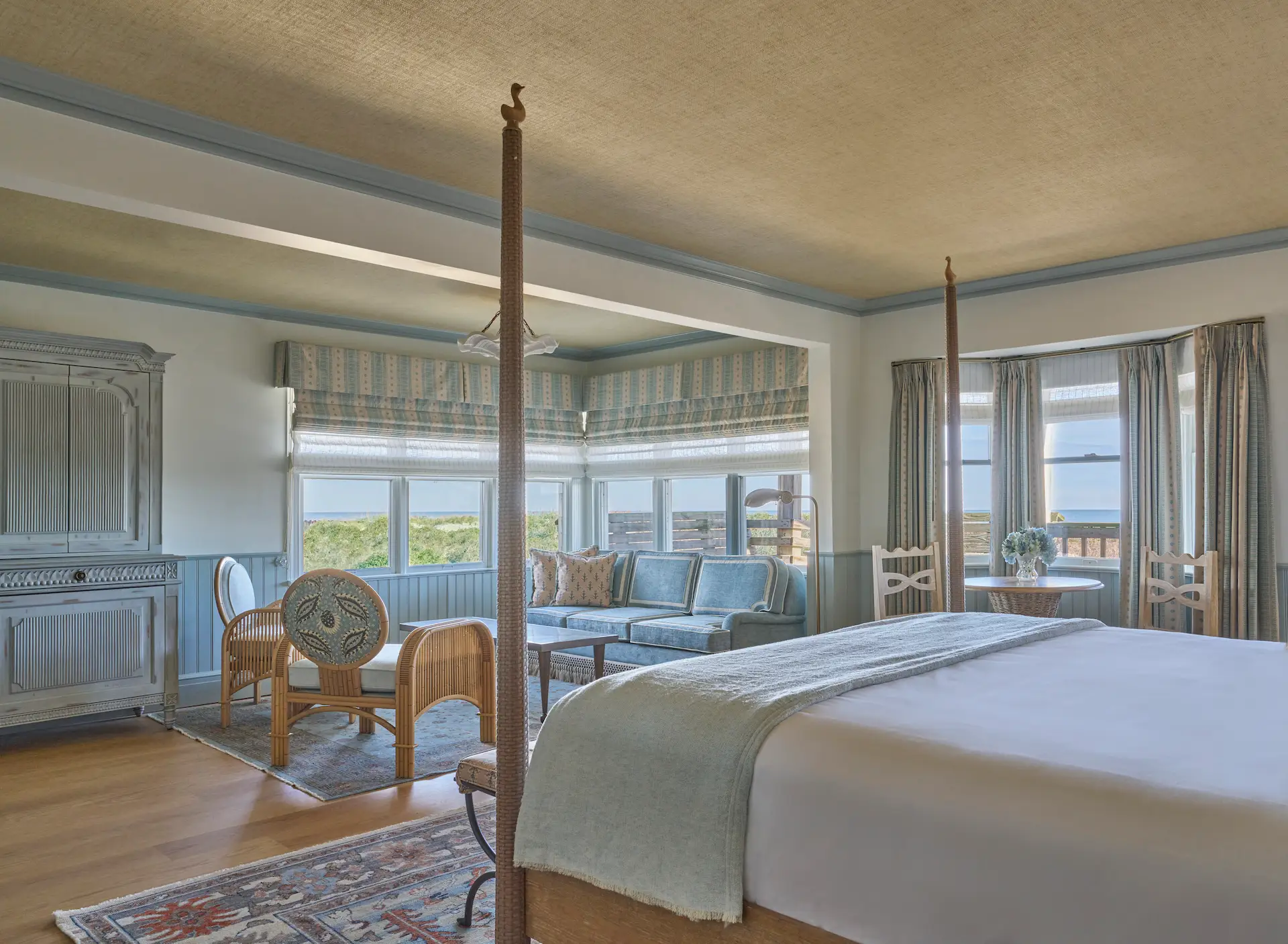 Spacious coastal bedroom with a four-poster bed, light blue sofa, wicker chair, and ocean views through large sunlit windows.