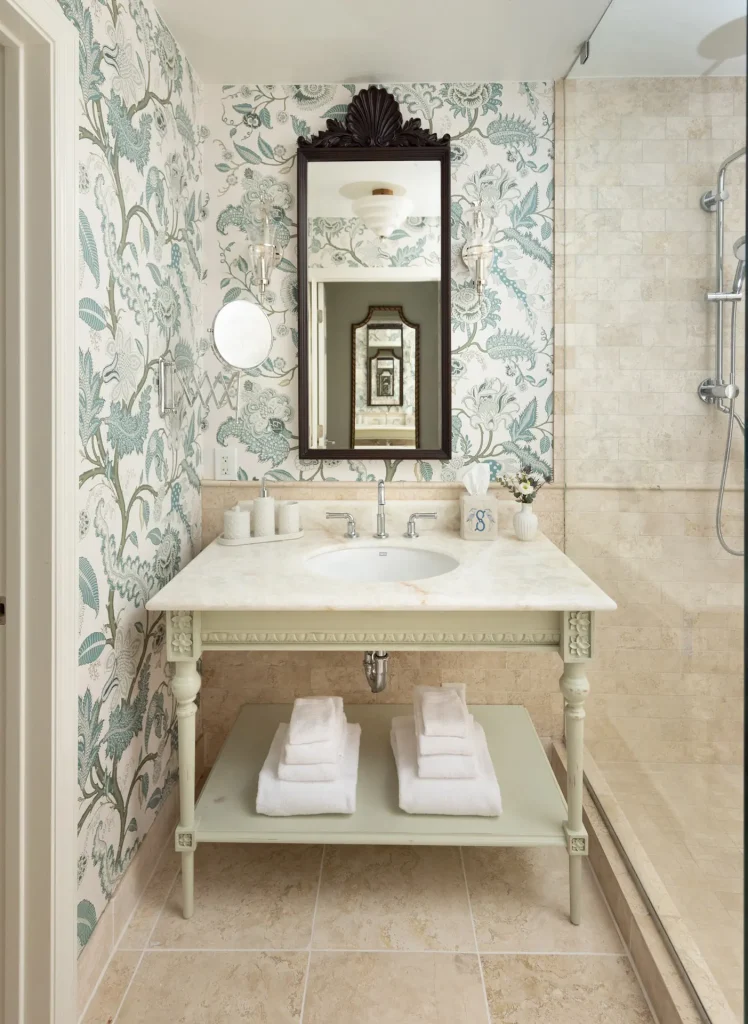 Vintage-style bathroom with floral wallpaper, carved mirror, and white vanity beside a beige stone walk-in shower.