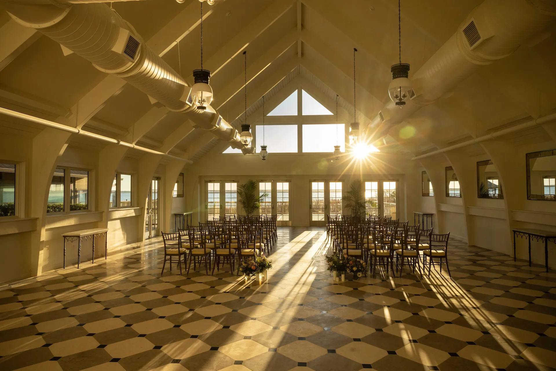 Elegant waterfront venue with vaulted ceilings, checkered tile floors, and round tables set for a sunlit celebration.