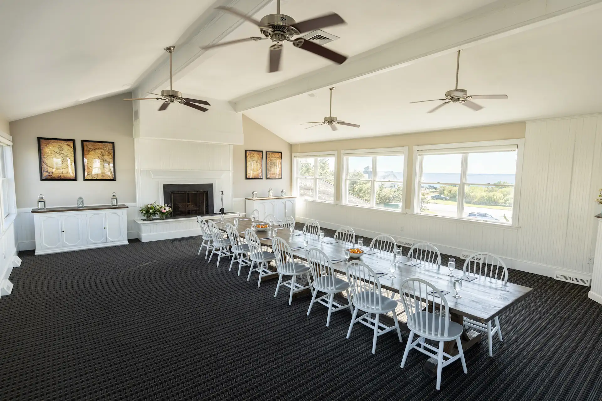Spacious, sunlit dining room with long table, white chairs, fireplace, ceiling fans, and windows overlooking landscape.