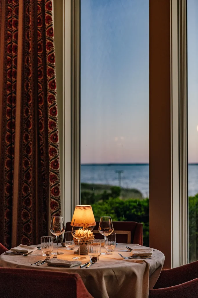 Elegant candlelit dining table set by a large window overlooking serene ocean at sunset.