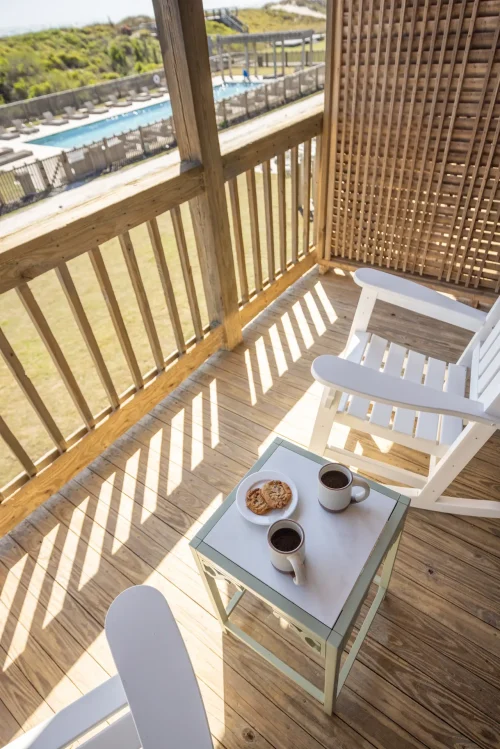 Two white rocking chairs and table with coffee and cookies on a sunny wooden balcony overlooking pool.