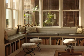 Bright reading nook with window bench, decorative pillows, three round stools, and shelves filled with books.