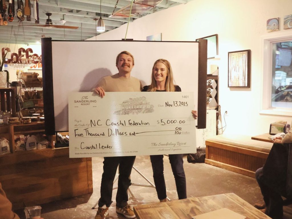 Two people indoors holding a $5,000 check to NC Coastal Federation in a cozy coffee shop setting.