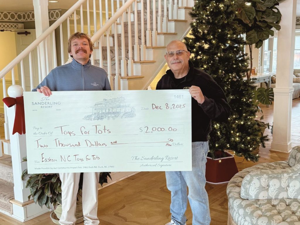 Two people indoors holding a $2,000 check to Toys for Tots beside a Christmas tree and staircase.
