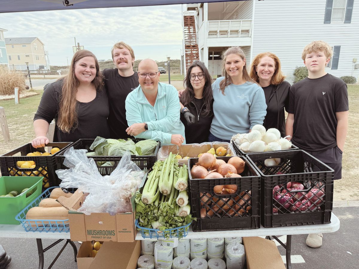 Smiling group at outdoor market table with fresh vegetables, onions, squash, and mushrooms.