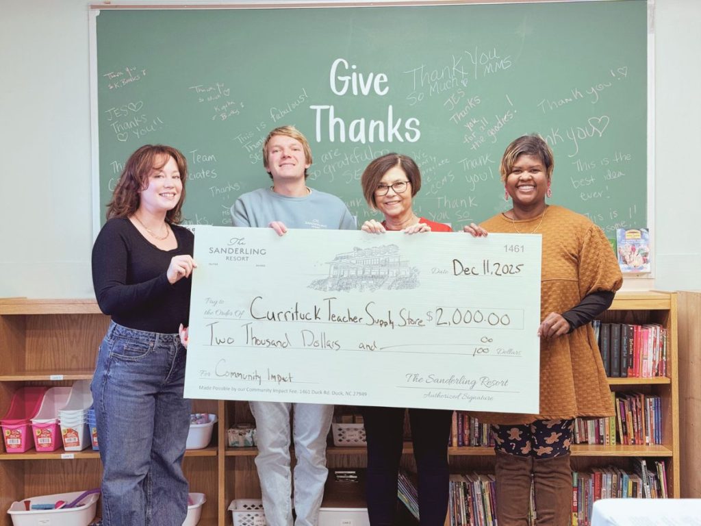 Four people holding a $2,000 check to Currituck Teacher Supply Store in front of a 'Give Thanks' board.