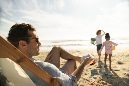 Relaxed man reading on a sunny beach while children play near the shoreline