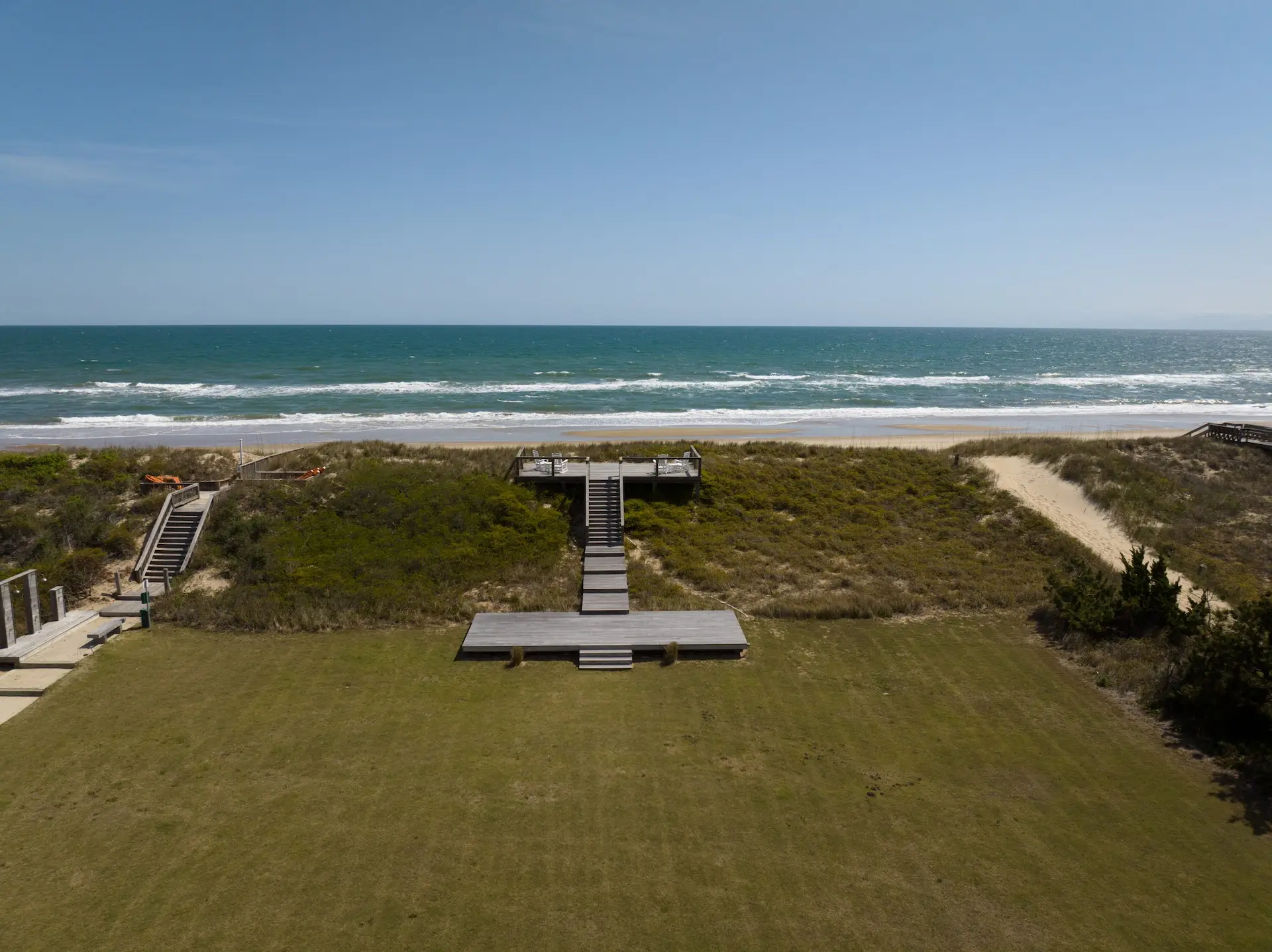 Wooden boardwalk and stairs lead to ocean-view deck with white chairs among coastal dunes.